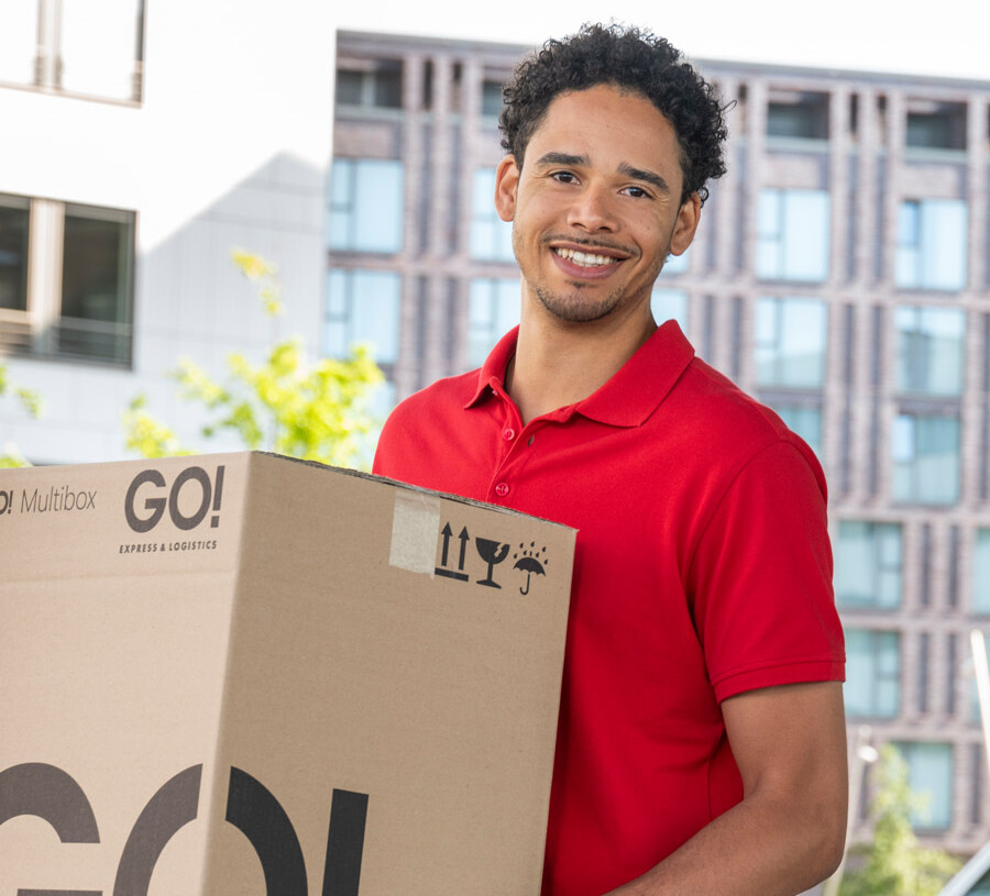 Lächelnder GO! Kurier mit mittelgroßem GO! Paket in der Hand vor einem Bürogebäude